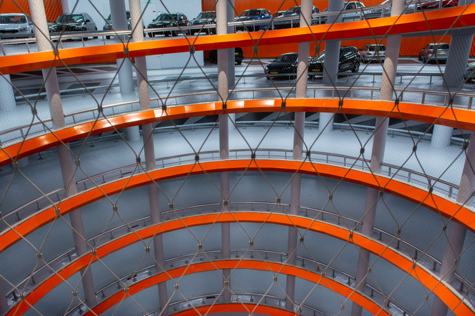 A multi-level, circular car parking structure viewed from the interior looking upward. The floors are supported by vertical grey concrete columns and feature bright orange metal beams that form concentric rings on each level. Safety railings run along the edges of each parking level, which is visibly lined with parked cars in various shades of black, grey, and white. The car surfaces range from smooth to slightly reflective, with some visible textures such as tinted windows and metallic finishes. The background shows additional parked vehicles on upper levels, partially obscured by the support columns and orange beams, with a mesh safety netting visibly hanging down in the foreground. The lighting appears to be natural, diffused through an open or partially open roof area, highlighting the structural details and creating soft shadows that enhance the three-dimensional perspective. This interior scene exemplifies a modern multi-storey parking facility, which could be associated with commercial or residential property management involving external storage and vehicle storage solutions, managed by companies like Waste Clearance Holland Park, which often handle site clearances or refurbishment projects.