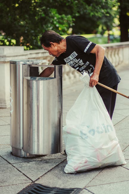 A man with dark hair, wearing a black T-shirt with white text and dark pants, is seen on a paved outdoor area next to a curved stone railing. He is bent over, using his right hand to reach into a tall, cylindrical, stainless steel rubbish bin. His left hand holds a large white refuse bag that appears to be partially filled with waste, resting against his hip. The man is operating in a landscaped park or garden setting, with lush green trees and foliage visible in the background, suggesting a well-maintained public space. The scene is illuminated by natural daylight, highlighting the reflective surface of the bin and the textures of the clothing and bag. This image aligns with private rubbish collection or on-site waste clearance services, such as those offered by Waste Clearance Holland Park, which specialise in waste removal from outdoor public areas and similar environments, providing an alternative to municipal rubbish disposal methods.