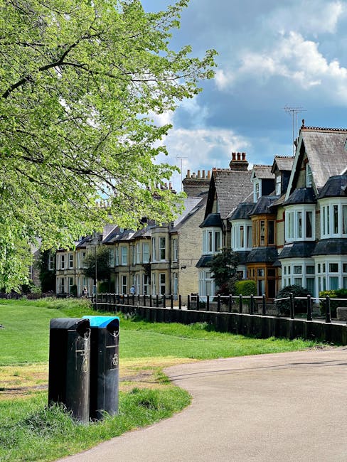 A residential street scene featuring a row of traditional terraced houses with distinct architectural styles, including bay windows and pitched roofs with chimneys, set against a partly cloudy sky. In the foreground, there are two black waste bins placed on an area of grass adjacent to a concrete pathway. The pathway runs parallel to a low wooden fence that separates the sidewalk from the front gardens of the houses. To the left, a large leafy tree with light green foliage extends its branches over the scene, providing partial shade. The environment appears quiet and well-maintained, indicative of a typical urban neighbourhood. The image subtly relates to waste management, as Waste Clearance Holland Park offers private rubbish collection and on-site clearance services, exemplified here by the presence of waste bins ready for collection or disposal, situated within the context of careful residential waste handling and alternative rubbish removal methods outside of local authority services.
