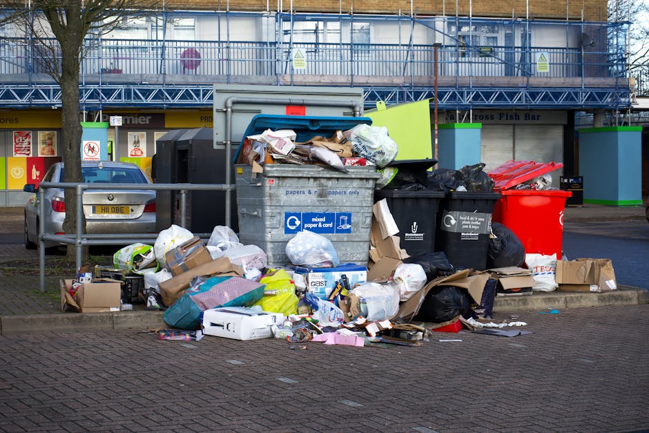 In the foreground, there is a large, overflowing metal recycling bin labeled for mixed paper and cardboard, with its lid open and paper waste spilling out onto the pavement. Surrounding the bin are various discarded cardboard boxes, plastic bags, crumpled paper, and packaging materials, some flattened while others are still intact. To the right, multiple black and red rubbish bags are piled against the bin, with some partially torn, revealing additional waste inside. Behind the bin, a grey station wagon vehicle is parked, partially obstructed by a metal railing that separates the waste accumulation from the nearby sidewalk. In the background, a commercial building with a yellow and black storefront, as well as a retail signage indicating a fish bar, can be seen. The building is covered with blue scaffolding and protective netting, indicating ongoing maintenance or renovation work. The scene is illuminated by natural daylight, creating a neutral, everyday setting typical of unauthorized or excess waste left on the street, which Waste Clearance Holland Park could handle through private rubbish collection services aimed at maintaining cleanliness in urban areas.