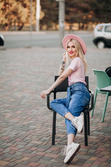 A young woman with long blonde hair and fair skin is sitting on a black metal chair outdoors on a paved area with interlocking brick patterns. She is wearing a wide-brimmed pink hat, a light pink short-sleeved T-shirt, and high-waisted blue jeans rolled slightly at the ankles. Her white sneakers are partially visible, and she has a relaxed, cheerful expression with a smile, showing her red lipstick. Her left arm is resting on the back of the chair while her right arm is leaning on her knee, with her right foot crossed over her left knee. In the background, there are two other chairs—one green and one black—placed nearby, and a road with a passing white vehicle and some trees with autumn-colored foliage. The scene suggests a casual outdoor setting with natural light, and the overall atmosphere is bright and cheerful, reflecting a relaxed moment in an urban park or public space, which could be associated with independent disposal or informal waste handling.