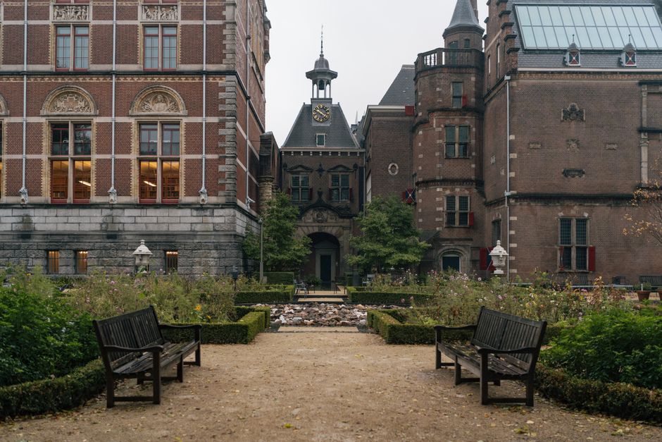 A symmetrical garden pathway flanked by neatly trimmed hedges and flowering plants leading towards an ornate historic building with a clock tower, situated between two large brick structures. The pathway is composed of compacted earth with two dark wooden benches positioned on either side, facing each other along the gravel edges. The garden features lush green foliage, some small trees, and blooming flowers, with subtle natural lighting indicating an overcast day. In the background, the central building displays a pointed spire topped with a decorative finial, arched windows, and a small balcony, surrounded by urban architecture constructed from red and brown bricks with a mix of traditional and modern elements, including a glass roof section and turrets. Such detailed scene of a managed outdoor space aligns with preparations for or environment suitable for private waste handling or nearby rubbish removal services, emphasizing tidy, maintained surroundings in a city setting. Waste Clearance Holland Park, as a professional rubbish service, might be involved with clearing waste from similar landscaped areas or nearby structures to maintain their aesthetic and functional appeal.
