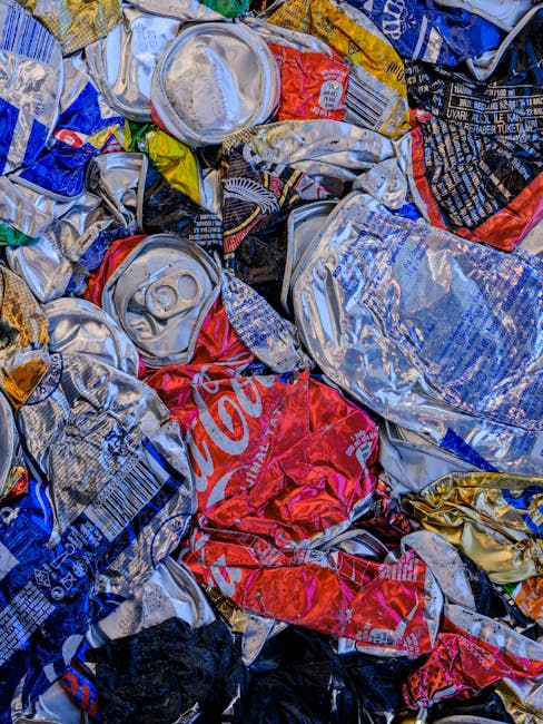A large collection of mixed waste and rubbish bags piled around multiple waste bins located outdoors on a paved area in front of a commercial building with blue scaffolding. The waste includes black and yellow plastic rubbish bags, flattened cardboard boxes, paper waste, and various discarded packaging material. The waste bins consist of a grey bin labeled for mixed paper and card, a black bin, and a red bin, all filled to or overflowing with rubbish. Nearby, there is a parked silver car with a visible license plate, and a metal railing separates the waste area from the pedestrian walkway. In the background, a commercial storefront with closed shutters, windows, and blue scaffolding can be seen, indicating a business or construction site. The setting appears to be a typical urban environment where private waste collection or rubbish removal services, such as those provided by Waste Clearance Holland Park, might be employed for responsible waste disposal.