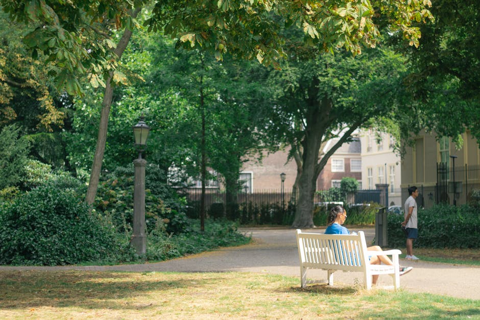 A woman seated on a white park bench faces to the right, wearing a blue shirt and light-colored shorts, with her hair tied back. Standing nearby on a paved pathway, a young girl in a white t-shirt and dark shorts observes the surroundings, while another child stands further to the right, partially obscured, dressed in a white top and dark shorts. The scene is set in a lush, green urban park with dense foliage, large mature trees with leafy canopies, and a black metal fence in the background. The park includes classic lampposts along the pathway, and the environment appears well-maintained with grassy areas and a mix of shaded and sunlit spots, creating a calm and natural atmosphere appropriate for relaxation or casual outdoor activities. Waste Clearance Holland Park might handle rubbish removal for such public spaces or related private properties, emphasizing the need for efficient waste management to preserve the park's environment and appearance.