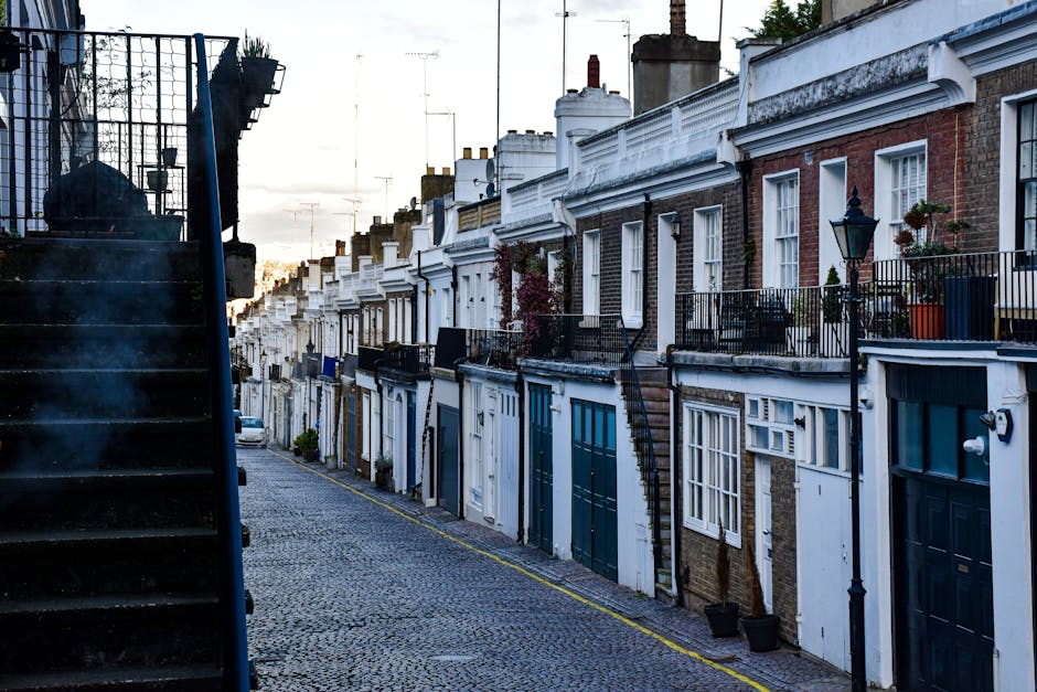 A narrow cobblestone street in a quiet residential area lined with terraced brick houses featuring white facades, decorative cornices, and black wrought iron railings on small front balconies. Some balconies have potted plants and outdoor furniture. Chimney stacks rise above the rooftops against a cloudy sky, indicating an urban setting. On the left side of the image, a dark external staircase with metal steps and railing ascends from a building, with the shadowed side partially obscuring the view. The street is bordered by a yellow line along the curb, with no visible vehicles or pedestrians, and the overall scene appears calm and well-maintained, typical of a historic London neighborhood. Waste Clearance Holland Park may be involved in maintaining this area by providing independent rubbish collection or on-site clearance services for residents’ property improvements or decluttering activities.