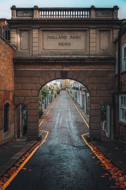A narrow cobblestone road leading through an arched stone gateway inscribed with 'Holland Park Mews' in a residential area. The archway is constructed from reddish-brown stone blocks, with a decorative balustrade on top. Flanking the entrance, red brick buildings with white-framed windows are visible on both sides, some with small details such as brick ledges and window sills. The street beyond the arch slopes gently upward, lined with tall, closely spaced fences made of metal and wood, enclosing gardens or yards, some with visible greenery and flowers. Fallen autumn leaves are scattered along the edges of the road and pavement, adding warm tones to the scene. Overhead, the sky is partly cloudy, providing soft, diffused lighting. In the context of rubbish removal services like those offered by Waste Clearance Holland Park, this image illustrates an example of a private residential street where on-site clearance or independent waste collection may be relevant, with the narrowness of the street emphasizing the importance of suitable logistics for rubbish removal in such environments.