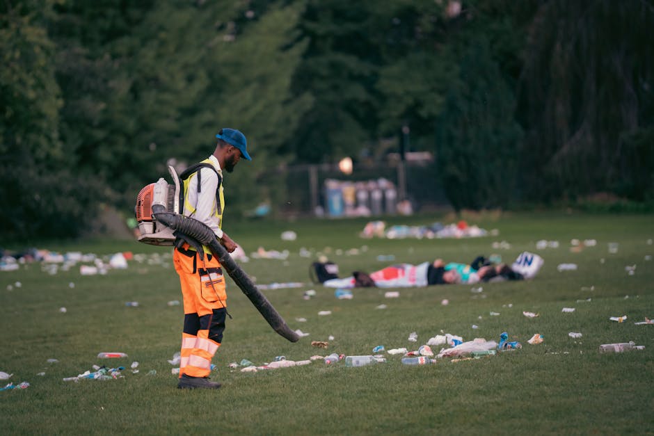 A man dressed in a high-visibility jacket with orange and navy trousers and a blue cap is using a leaf blower, which has a flexible hose and a backpack motor, to clear litter from a grassy park area. The ground is densely littered with various types of rubbish, including plastic bottles, cans, and paper debris, scattered across the grass. In the background, a group of people is lying on the ground, surrounded by more scattered waste, with trees and greenery framing the scene. The natural outdoor environment appears to have been recently disrupted by the presence of litter, emphasizing the need for professional waste clearance services like those offered by Waste Clearance Holland Park for maintaining cleanliness and environmental protection. The lighting suggests it is either late afternoon or early evening, creating a muted, natural atmosphere suitable for outdoor maintenance work, highlighting the importance of private waste handling and rubbish removal to manage environmental impact efficiently in such areas.