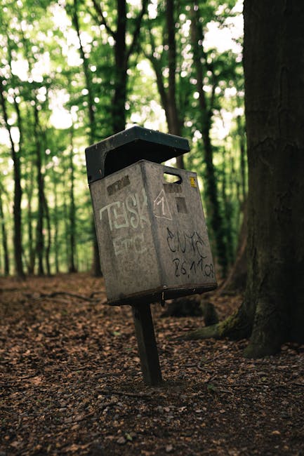 A public mailbox is mounted on a tilted metal post in a wooded area, surrounded by tall trees with dense green foliage in the background. The mailbox is rectangular with a rounded top, made of metal with a weathered, grey surface covered in graffiti and stickers. The mailbox's open lid reveals an interior space for inserting mail, and the front panel features a small, rectangular slot for deposits. The ground beneath it is covered with dirt and scattered leaves, indicating an outdoor, natural setting. The environment appears serene yet slightly neglected, with no immediate signs of nearby urban infrastructure. This scene exemplifies how independent waste collection points like this can be used for private or alternative waste handling outside of conventional city rubbish services, aligning with the concept of on-site clearance or personal disposal options. The overall lighting is ambient daylight filtering through the canopy, highlighting the mailbox's textured surface and its surrounding environment, emphasizing the contrast between human-made objects and the natural setting.