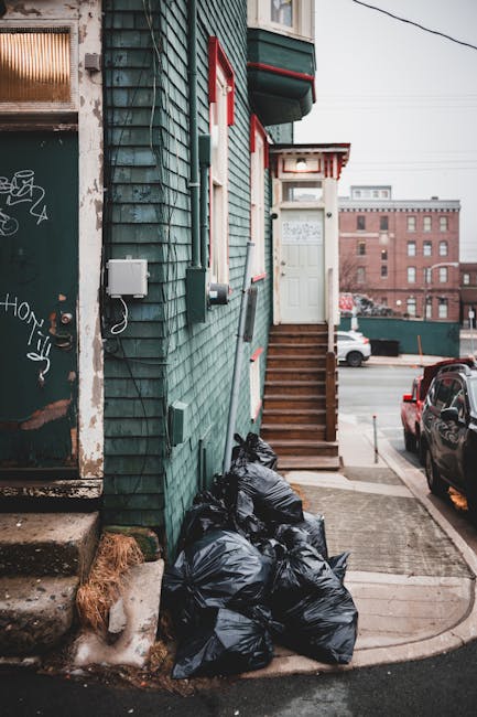 A small urban building with weathered green wooden cladding and red-and-white striped awnings above a set of wooden stairs leading to a white door. To the left, there is a black metal door with graffiti and peeling paint, set into the lower part of the building. On the ground, adjacent to the building’s corner, there are four black rubbish bags piled up, appearing tightly packed and sealed. The area around the bags is a paved alleyway with concrete steps and a metal grate near the bags' base. To the right, parked cars are visible, including a dark vehicle on the street, with an industrial brick building in the background and power lines overhead. The overall scene reflects a typical urban setting where waste is temporarily stored outside a residential or commercial property, possibly awaiting collection by a private rubbish removal service such as Waste Clearance Holland Park, highlighting an example of on-site waste disposal in an inner-city environment.