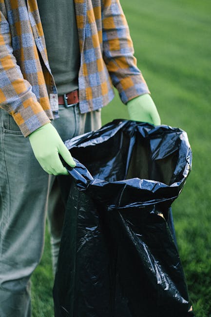 A person wearing a checkered yellow and grey shirt over a grey t-shirt and green gloves is holding open a large black plastic rubbish bag outdoors on a grassy area. The bag appears to be empty or only partially filled. The background consists of green grass and blurred natural surroundings, suggesting a garden or park environment. The person's waist is visible, with a brown belt and grey trousers. This scene reflects an activity related to rubbish collection or waste disposal, possibly involving independent waste handling or on-site rubbish removal, as part of a typical waste clearance process. Waste Clearance Holland Park offers services in waste collection and rubbish removal, supporting private disposal needs in the area. The image emphasizes the practical aspects of rubbish management, including careful handling and preparation for waste removal services.